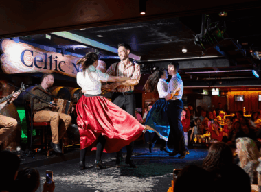 Traditional Irish dancers perform energetically on stage at a vibrant, bustling bar.