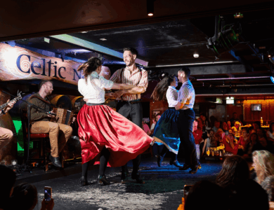 Traditional Irish dancers perform energetically on stage at a vibrant, bustling bar.