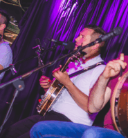 Three musicians perform live at the Arlington Hotel bar with guitars and drums.