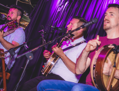 Three musicians perform live at the Arlington Hotel bar with guitars and drums.