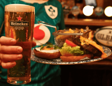 Person in rugby shirt holds a Heineken beer and a plate with a burger and chips at a cosy bar.