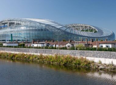 Modern stadium near riverside houses in Dublin, sunny day scene.