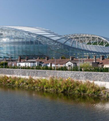 Modern stadium near riverside houses in Dublin, sunny day scene.