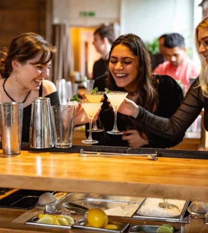 Three women laughing and clinking cocktails at a lively bar.