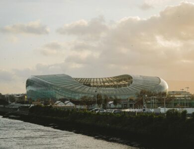 Modern stadium near waterside at sunset, clouds and cityscape in the background.