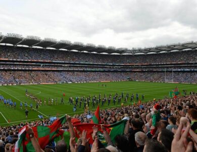 Crowds cheer a match at Croke Park, colourful flags wave, and teams line up on the pitch.