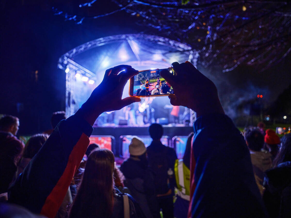 People enjoy a live outdoor concert, capturing moments on their phones.