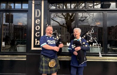 Two people in blue shirts play bagpipes outside a bar and restaurant in Dublin.