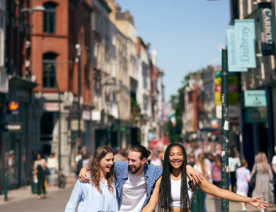 Three friends walk joyfully on a bustling Dublin street on a sunny day.
