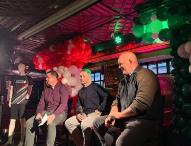 Three men seated on stage with microphones, surrounded by colourful balloons.