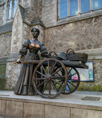 Bronze statue of a Victorian fishwife pushing a wooden cart with baskets outside a stone city church in Dublin