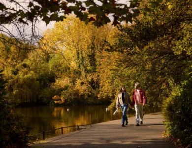 Couple strolling beside a tranquil tree-lined lake in autumn, warm hotel getaway atmosphere, visible text: "Louis Fitzgerald Hotel" not shown in image