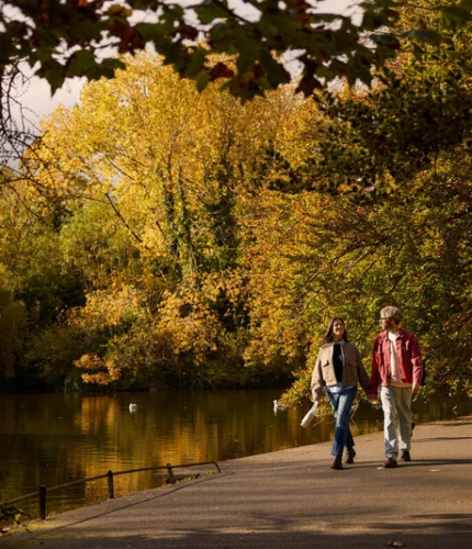 Couple strolling beside a tranquil tree-lined lake in autumn, warm hotel getaway atmosphere, visible text: "Louis Fitzgerald Hotel" not shown in image