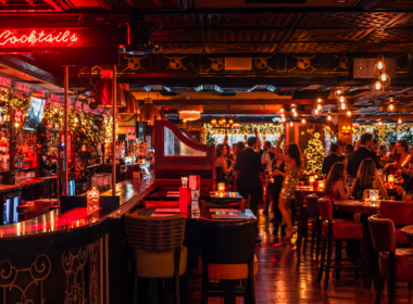 Festive hotel bar with warm lighting, guests chatting and dancing near the cocktails sign, Christmas tree visible